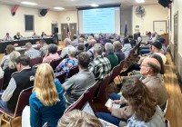 A packed city council meeting showing crowd, projection screen, and council members.