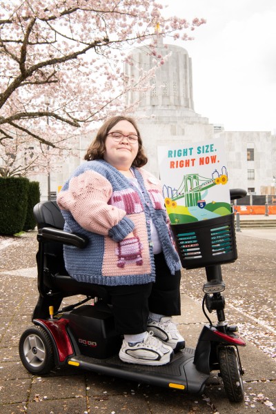 Cassie sits on a mobility device facing the camera, with a poster that shows a bridge and says "Right size. Right now." She is in front of the Salem capitol.