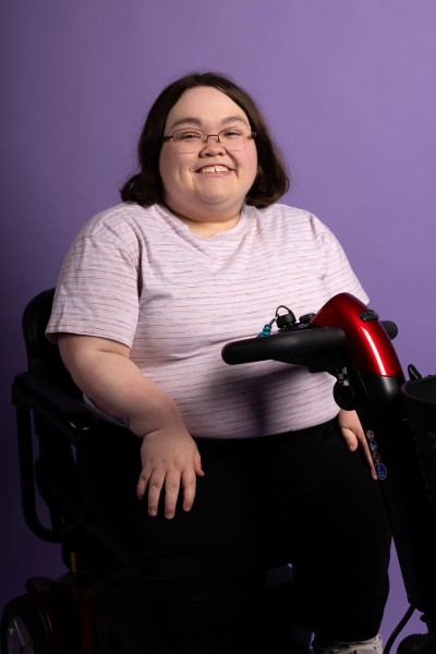 Cassie Wilson sits on an assistive mobility device against a purple background and faces the camera, smiling