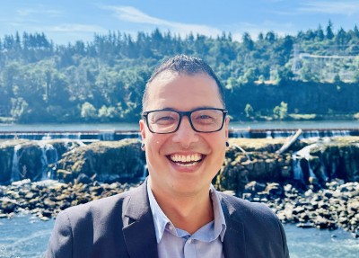 Justin stands outside with Multnomah Falls in the background and smiles at the camera on a sunny day