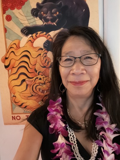 Anita faces the camera and gently smiles. She is wearing a purple lei and a piece of art hangs behind her.