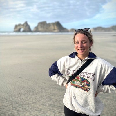 Anna stands on a beach while wearing a sweatshirt and smiles at the camera