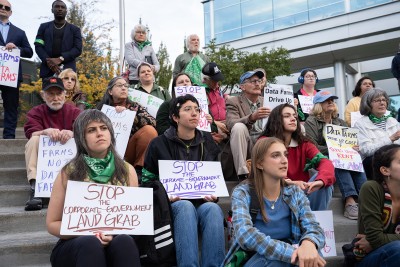 A group of people sit outside on steps and holds signs that read "stop the corporate-government land grab"