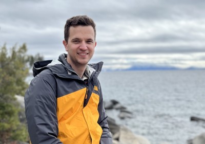 John sits outside on a rocky shore and smiles at the camera