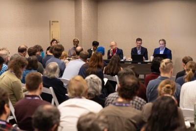 A group of panelists seated at a long table in front of a large crowd.