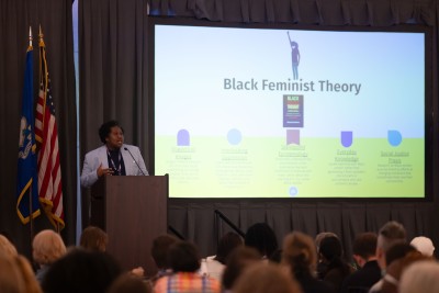A woman standing at a lectern in front of a crowd of people, with a projected presentation on a white screen that says, "Black feminist theory."