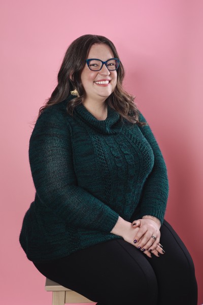 A smiling woman with shoulder-length brown hair and blue glasses. She is wearing gold earrings and a green sweater.