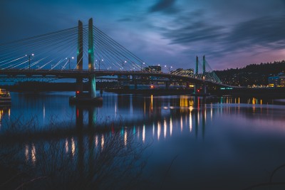 Tilikum Crossing, a cable-stayed bridge across the Willamette River, is shown at dusk with lighted reflections in the glassy surface of the river. In the background are hills with the lights of homes and vehicles glowing in the dark. The sky is blue and purple.