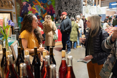 A crowded indoor market scene is framed by two people having a conversation in the foreground, and a countertop filled with bottles of wine
