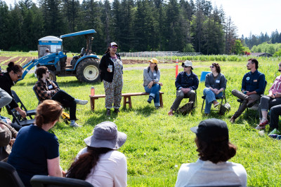 A group of people seated in chairs arranged in a circle in a grassy field near rows of agricultural crops.