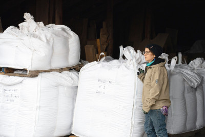 A person with a tan jacket, blue jeans, and a dark beanie stands next to large (bigger than they are) bundles of seeds