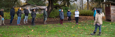 A person presenting to a group of people; they're all standing between a grove of oak trees and an agricultural field