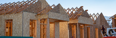Housing being built, with frames and plywood walls up. On the right side, a man on a ladder is hammering on one of the walls