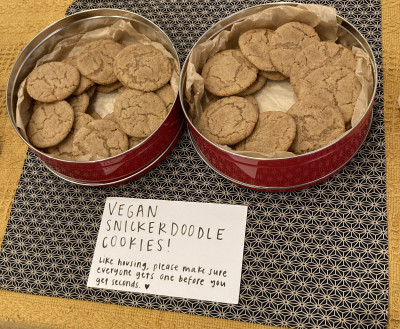Two tins of cookies with a handwritten sign that reads, "Vegan snickerdoodle cookies! Like housing, please make sure everyone gets one before you get seconds."