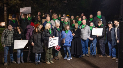 A group of people standing outside in the dark, with signs that say, "Keep Oregon Green," "Grow food not data," "Data centers are not your constituents, Sen. Sollman," and more. Many people are wearing green bandanas around their necks.