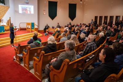 A packed church, with a woman in front presenting; the slide being projected on the wall is a Simpsons reference: "Hillsboro is being sold a monorail"