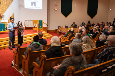 A man and a woman stand in front of a large crowd seated in wooden pews in a church