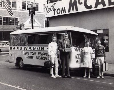 Tom McCall and his wife and kids standing by a bus painted with the slogan, "Band Wagon: Join your neighbors. Climb aboard!"