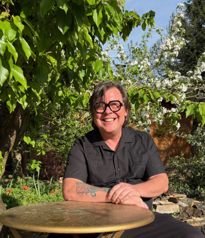 A smiling man with glasses, a navy blue shirt, and a tattoo on his arm in a sunlit garden