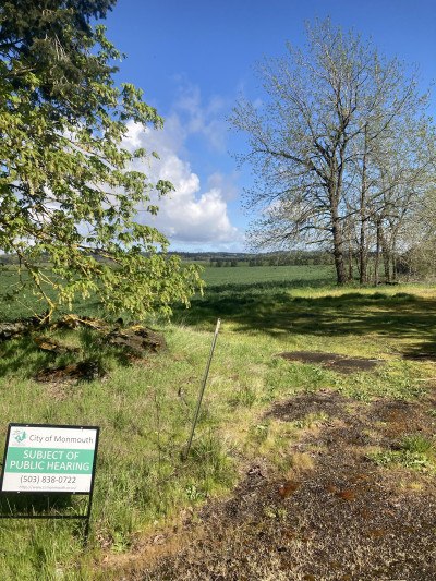 Forested and agricultural land with a sign in front of it: "City of Monmouth: Subject of Public Hearing"