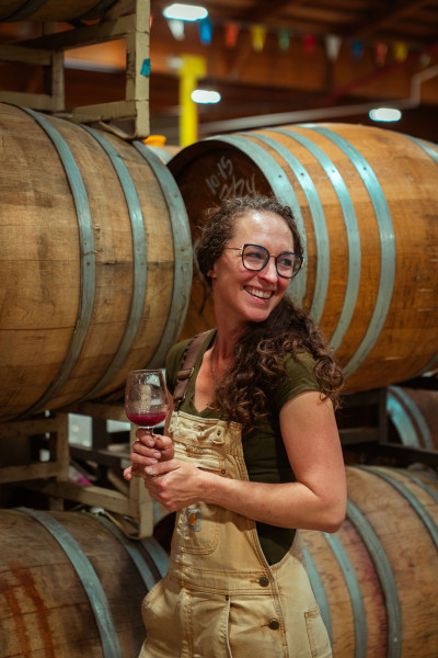 Meredith Bell smiles while holding a glass of red wine in a warehouse or large wine cellar, surrounded by large wooden barrels. She has long, curly brown hair; round glasses; and is wearing tan overalls over a dark green shirt, looking off to the side.