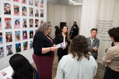 A group of people standing in a circle near a wall of colorful, photo portraits arranged in a grid on the wall