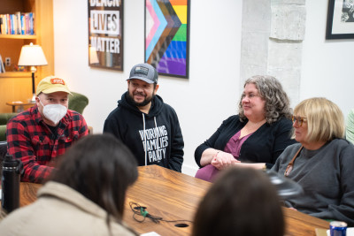 A group of people talking around a table in an indoor setting. There is a rainbow flag on the wall next to a Black Lives Matter banner.