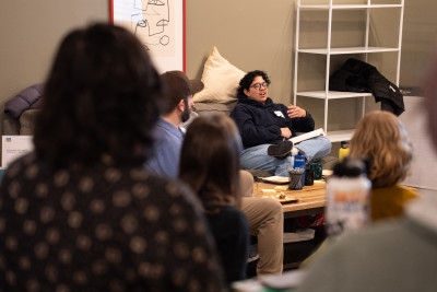 A person with short hair and a hoodie talks, while gesturing with their hands, as a group of people looks and listens.