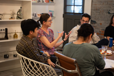 A woman in glasses and a purple shirt gestures with her hands while talking in a group setting, around a wooden table.