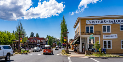 Main street with old-fashioned Western storefronts with bright paint, including the most prominent, a mustard-yellow building with the sign, "Sisters Saloon," in a classic American Western font. There are people walking on the sidewalks, and cars on the two-lane road.