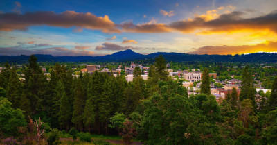 Aerial image of a city surrounded by trees and mountains, with a colorful sunset unfolding