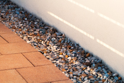Colorful river rock is arranged neatly in a border alongside a white exterior wall, and alongside a red brick walkway.