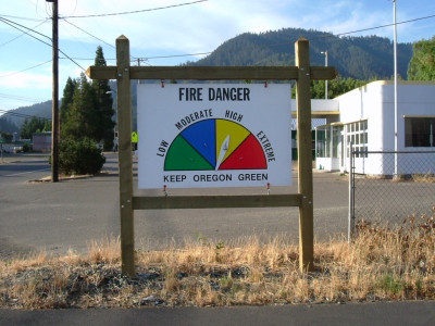 An outdoor sign in a large wooden stand, that reads, "Fire Danger. Keep Oregon Green." The chart ranges from low and moderate to high and extreme. The moveable arrow is currently pointing towards "high." The sign is set along a road in a small town with a prominent, forested hill behind it.