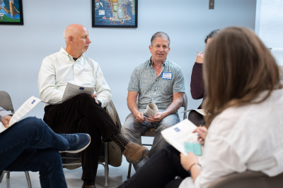 Five people sit on chairs arranged in a circle in an indoor setting, holding papers and wearing nametags. They appear to be talking to one another.