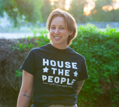 A smiling person wearing a shirt that says, "House the People--Welcome Home Coalition"