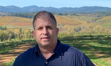 A person wearing a navy blue collared shirt faces the camera, while positioned in front of a scenic landscape of rolling farmland, fields in autumn colors, and distant forested hills under a clear blue sky.