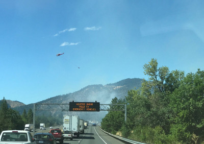 A backed-up freeway surrounded by lush vegetation on both sides, with hills partially obscured by smoke ahead. There is also a helicopter in the air and a signboard that reads, "Wildfire ahead. Yield to emergency vehicles."