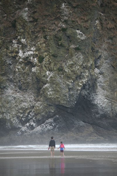 Two kids stand on wet sand near a giant sea stack, with a small, white-capped wave breaking near the rock's base. The rock takes up most of the frame.