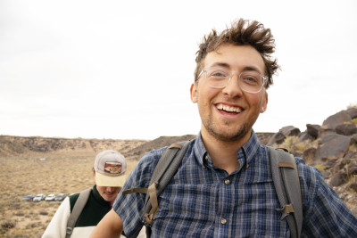 A smiling young man with glasses, a blue plaid shirt, and a backpack in an arid, rocky landscape.