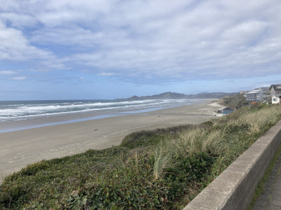 A wide, sandy beach framed by blue water on the left, beach grass and a low cement wall on the right. In the distance are buildings and a large headland with a lighthouse.