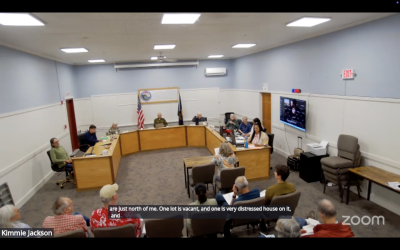 Elevated view of a conference room/council chamber where a person is testifying in front of 8 people at elevated seats/desks.