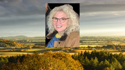 A photo of a smiling woman with light hair, tortoiseshell glasses, and a tan jacket, superimposed over a pastoral landscape