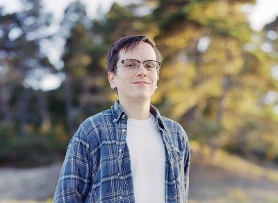 A man with black glasses, a blue flannel shirt, and a white t-shirt underneath, standing in an outdoor setting.