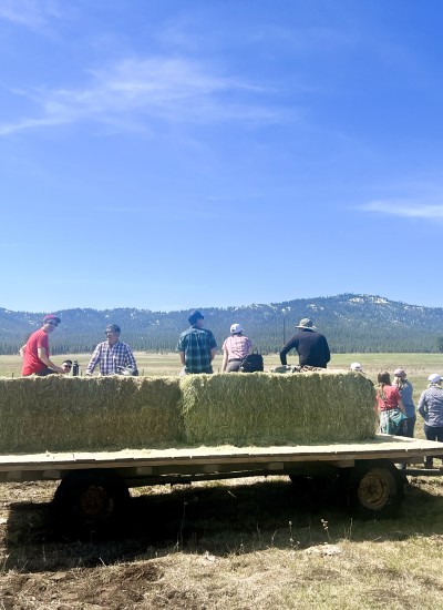 A group of people sit on bales of hay pulled by a tractor in a wide open field against a low mountain range and blue sky.
