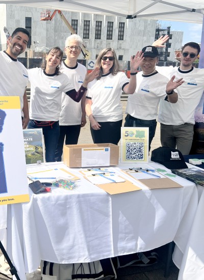 A group of 6 people smile and wave at the camera. They are standing at a booth in front of the capitol building in Salem.