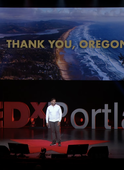 Sam Diaz stands alone on a dark stage with the words TEDxPortland behind him. On a screen, the words "thank you, Oregon" show atop a photo of the Oregon coast.