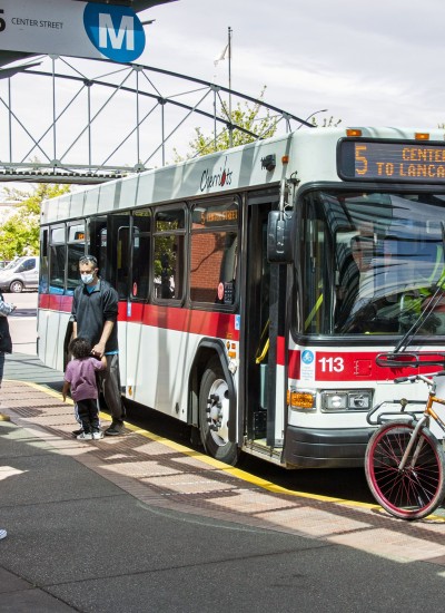 At a city bus stop, a person loads their bike onto the front of a bus while others stand and wait
