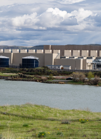 A large, windowless, industrial building alongside a wide river. There are arid hills on both sides of the river.