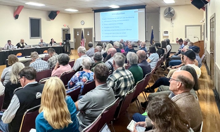 A packed city council meeting showing crowd, projection screen, and council members.
