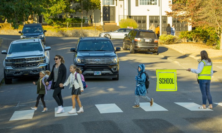 Children walk on a crosswalk with the help of an adult crossing guard. A car waits for them.